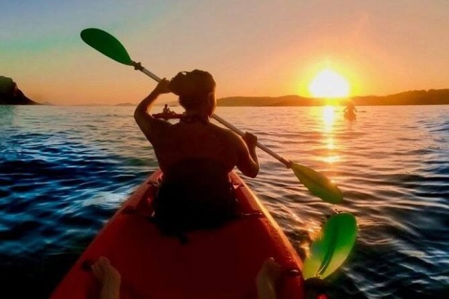 Person kayaking on calm water at sunset with mountains in the background.